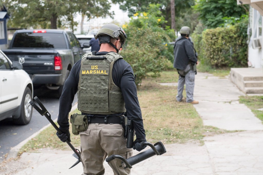 A U.S. Marshal in tactical gear, carrying tools, stands outside a house during Operation Triple Beam in San Antonio.