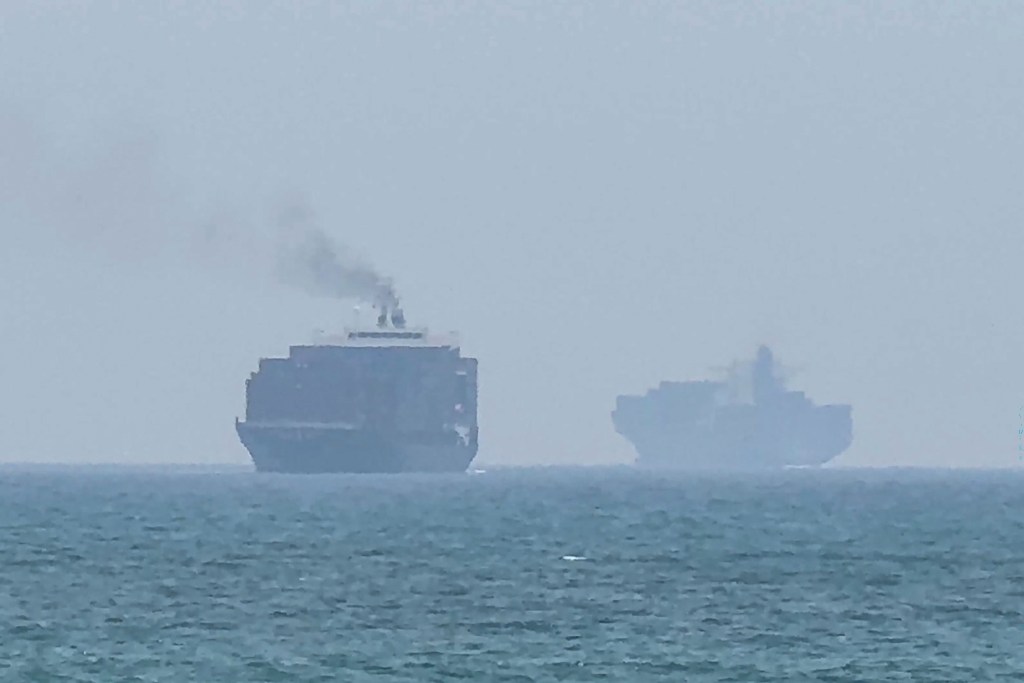 Two cargo ships on a hazy sea, one with smoke emanating from its top.
