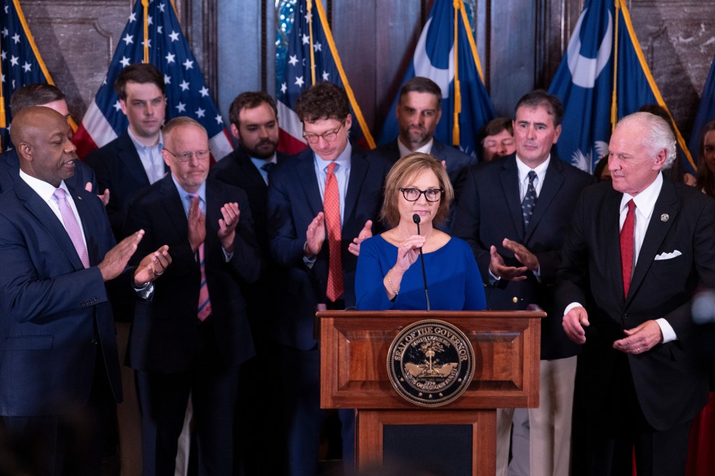 Darline Graham Nordone at a podium with South Carolina Gov. Henry McMaster and Sen. Tim Scott.