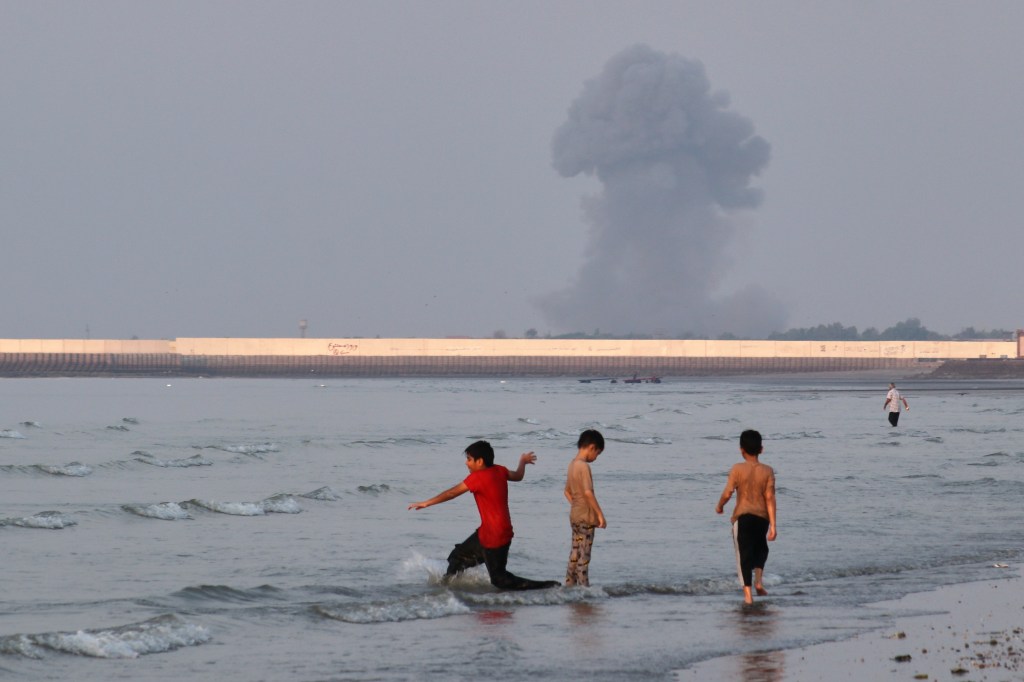 Three boys play in the shallow waters of the Strait of Hormuz, with a plume of smoke from an explosion rising in the background.
