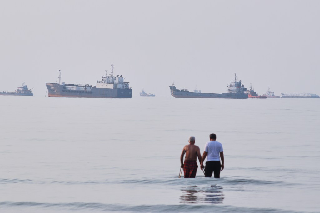 Two men wade in the waters of the Strait of Hormuz with vessels anchored in the background, off Bandar Abbas, Iran.